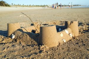 A sandcastle on the beach with small shells embedded in its walls, with a few people walking along the shoreline in the background.