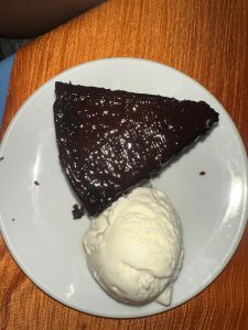 A slice of chocolate cake topped with chocolate glaze, accompanied by a scoop of vanilla ice cream on a white plate against an orange fabric background.

