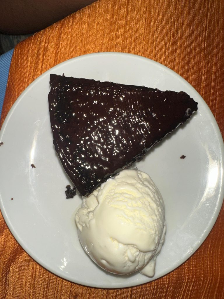 A slice of chocolate cake topped with chocolate glaze, accompanied by a scoop of vanilla ice cream on a white plate against an orange fabric background.