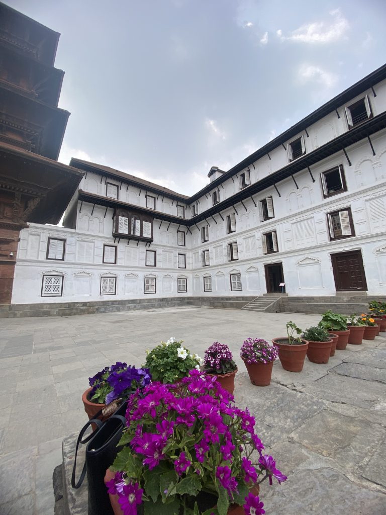 A courtyard with a historic building featuring white walls and numerous windows. In the foreground, a row of potted flowers adds vibrant colors, including purple and white blossoms, on a stone-paved surface under a cloudy sky.
