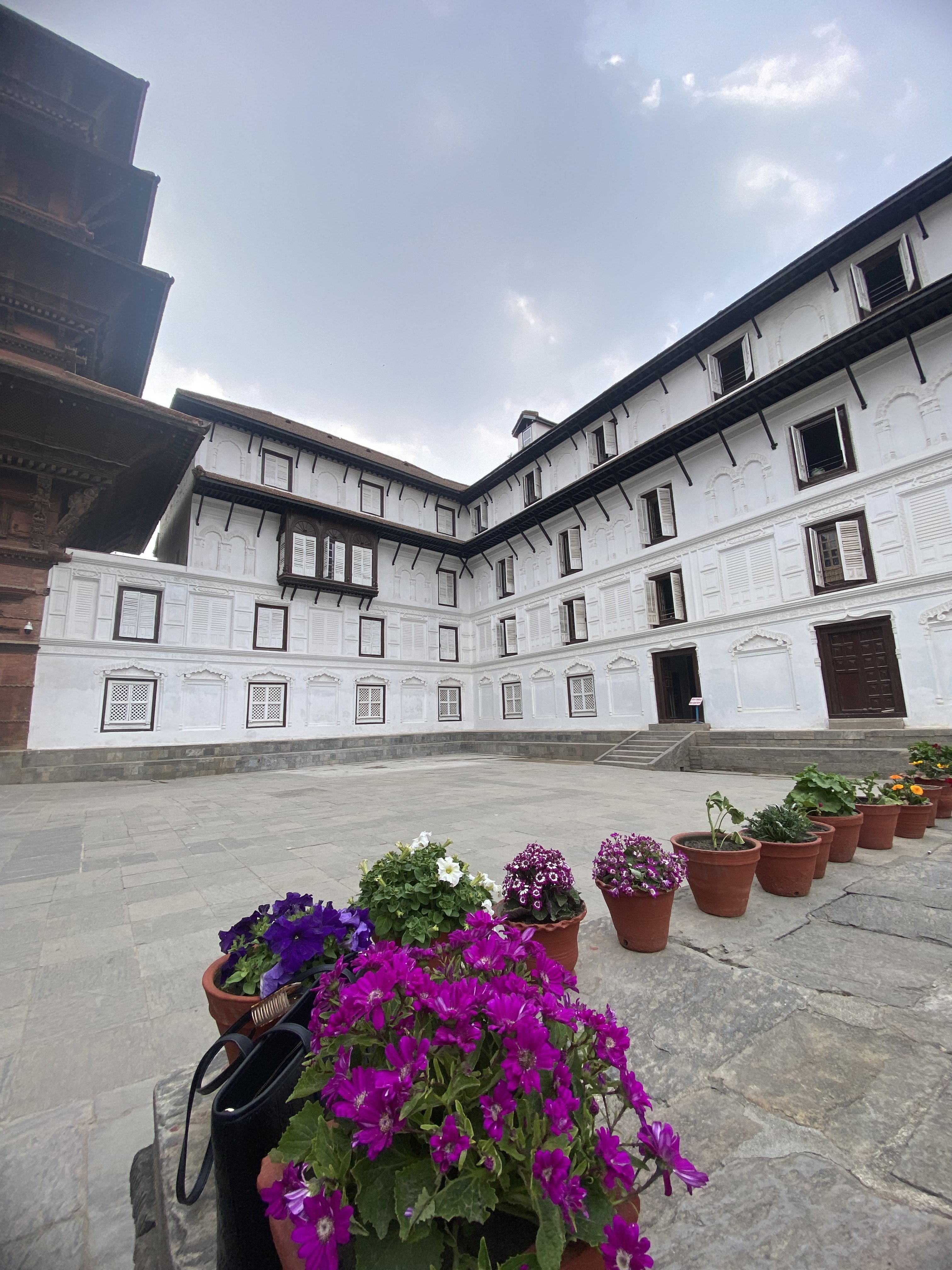 A courtyard with a historic building featuring white walls and numerous windows. In the foreground, a row of potted flowers adds vibrant colors, including purple and white blossoms, on a stone-paved surface under a cloudy sky.