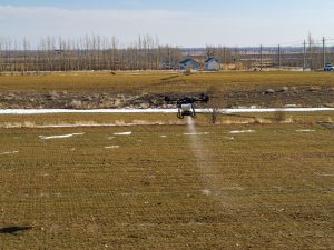 A drone flying over a grassy field, spraying a substance onto the ground. Beyond the field, there are sparse trees, a few small buildings, and power lines in the background under a clear sky.
