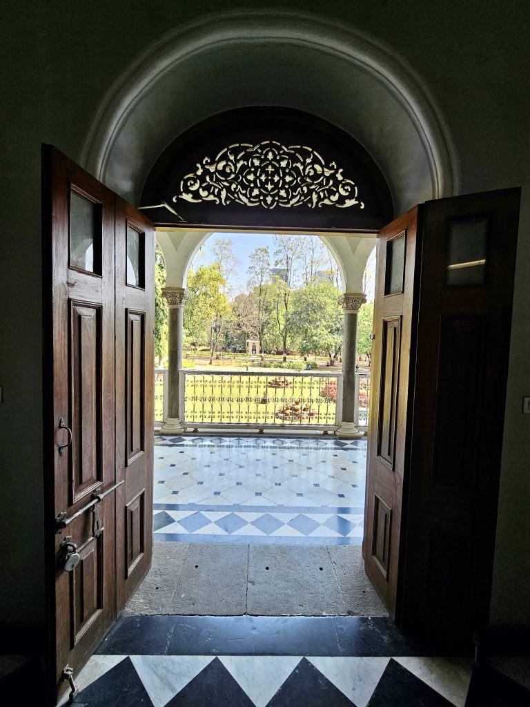 A doorway at the Aga Khan Palace in Pune, Maharashtra, leading to a garden area. The doorway features two wooden doors, a black and white tiled floor, and an arched transom with decorative latticework.