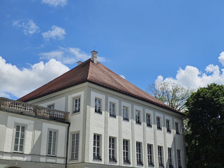 A portion of Nymphenburg Palace, Munich, features its white facade, rows of windows, and a red-tiled roof against a blue sky with scattered clouds. Part of the palace extends to the left, while trees are visible on the right.