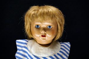 A close-up image of a creepy vintage doll with blonde hair, blue eyes, and a surprised expression, wearing a striped blue and white outfit with a white collar. 
