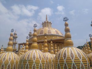 The Golden Mosque at Tangail, a large, ornately designed mosque featuring multiple domes and minarets with intricate mosaic patterns in golden and white colors. The domes are topped with decorative elements bearing Arabic script. The sky above is partly cloudy.
