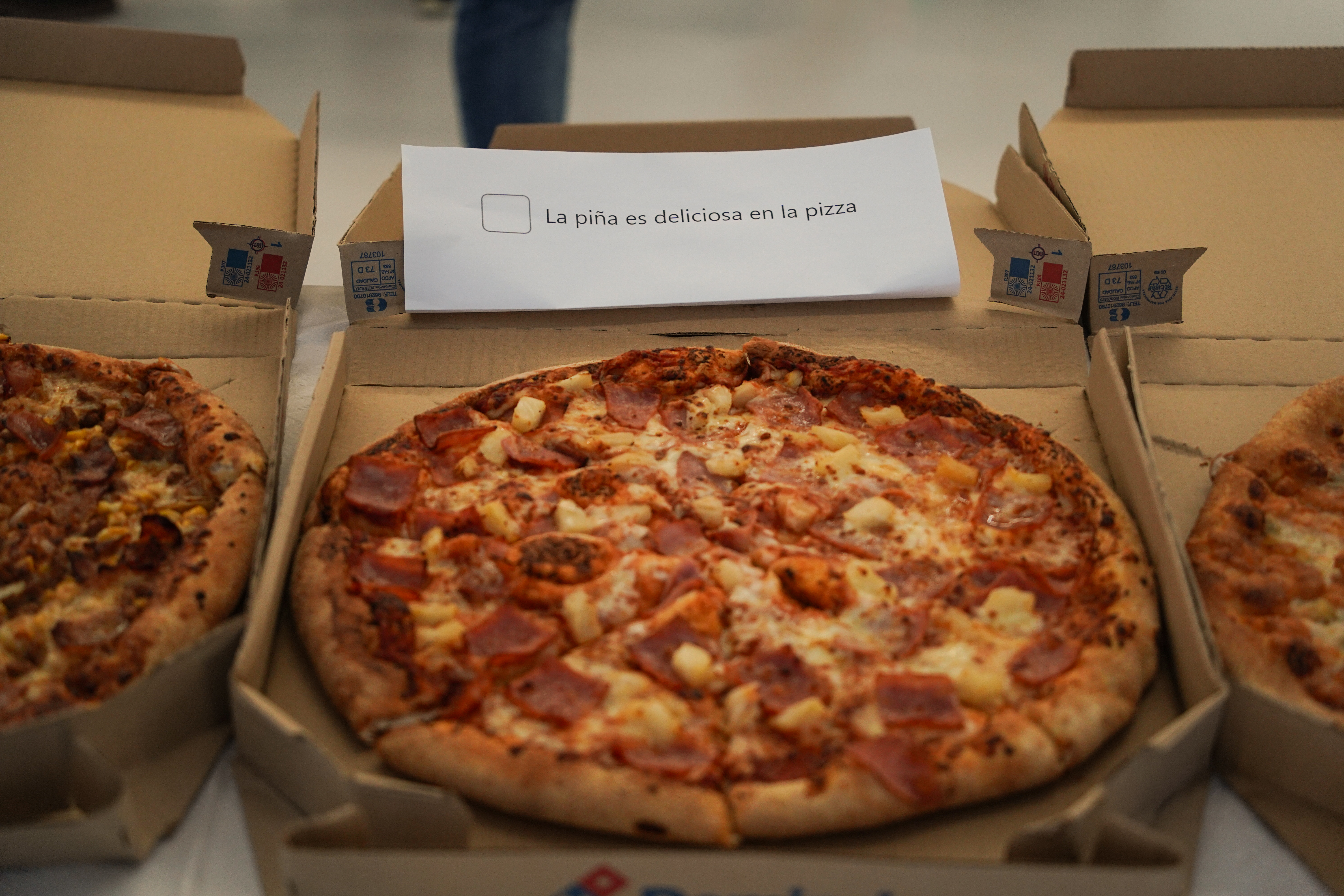 A variety of pizzas are displayed in open cardboard boxes. The pizza in the center has pineapple and ham toppings. A piece of paper above it reads "La piña es deliciosa en la pizza" which translates to "Pineapple is delicious on pizza."