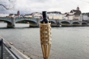 A bamboo torch in focus, with a blurred background of a river and a stone bridge, along with a cityscape and cloudy sky.