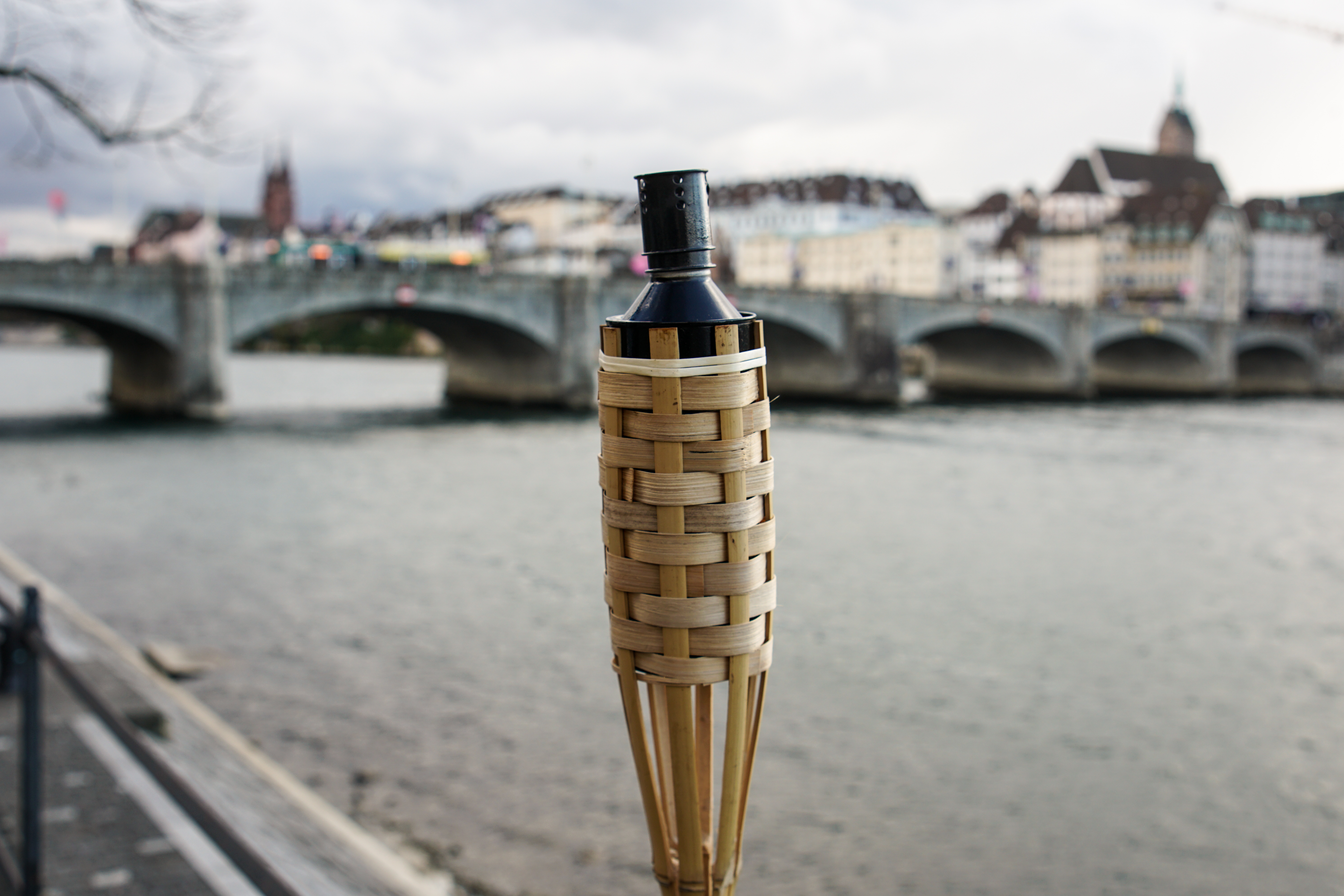 A bamboo torch in focus, with a blurred background of a river and a stone bridge, along with a cityscape and cloudy sky.