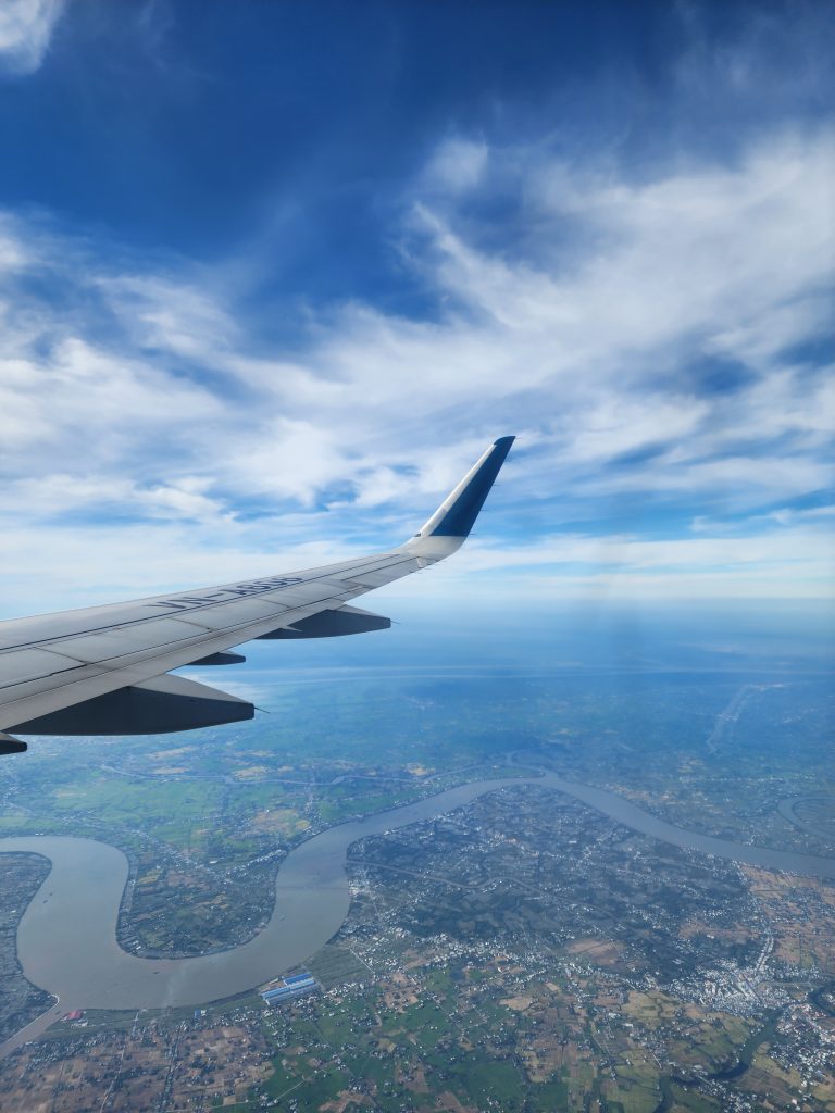 Aerial view of Ho Chi Minh City, Vietnam, from an airplane window. The airplane wing is visible in the foreground, with a winding river and cityscape below. The sky is bright blue with scattered white clouds.