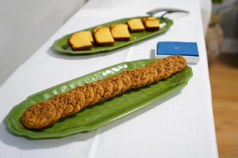 A long green platter with a row of chocolate chip cookies on a table, with a second green platter in the background holding sliced pound cake.