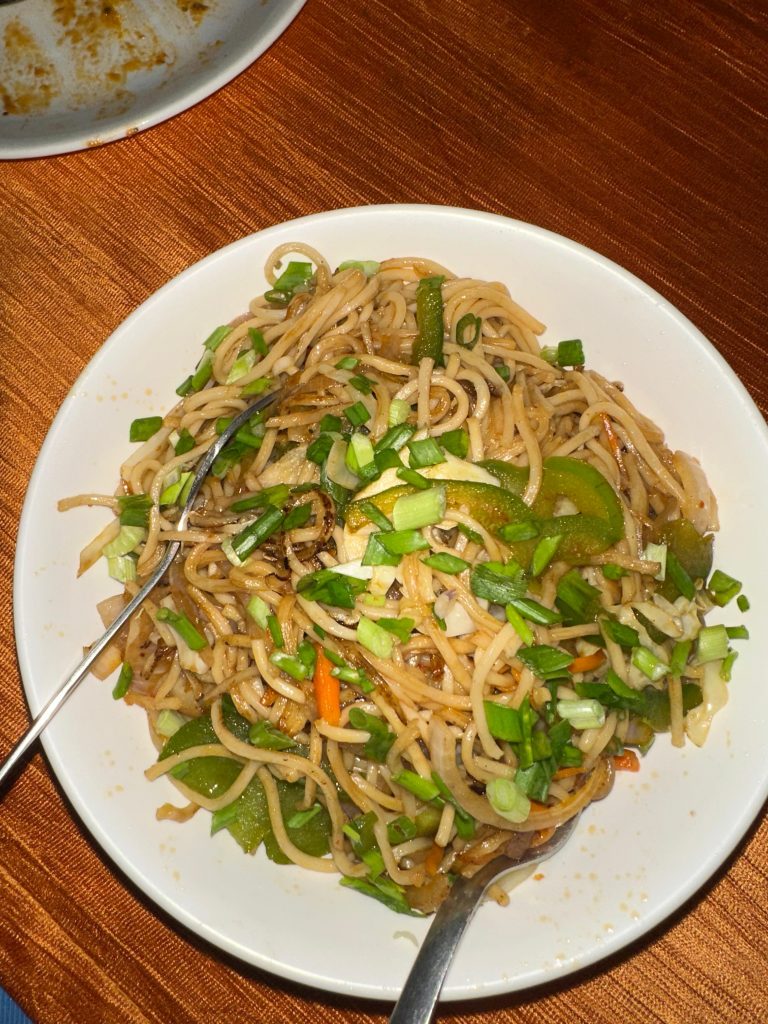 A plate of chowmein with green bell peppers, carrots, and chopped spring onions on top, set on a textured, orange tablecloth. Two utensils are placed in the noodles.