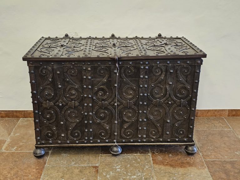 An antique, dark metal chest with ornate designs and studded detailing. It features a hinged lid and rests on four spherical feet, Hohensalzburg Fortress, Salzburg.