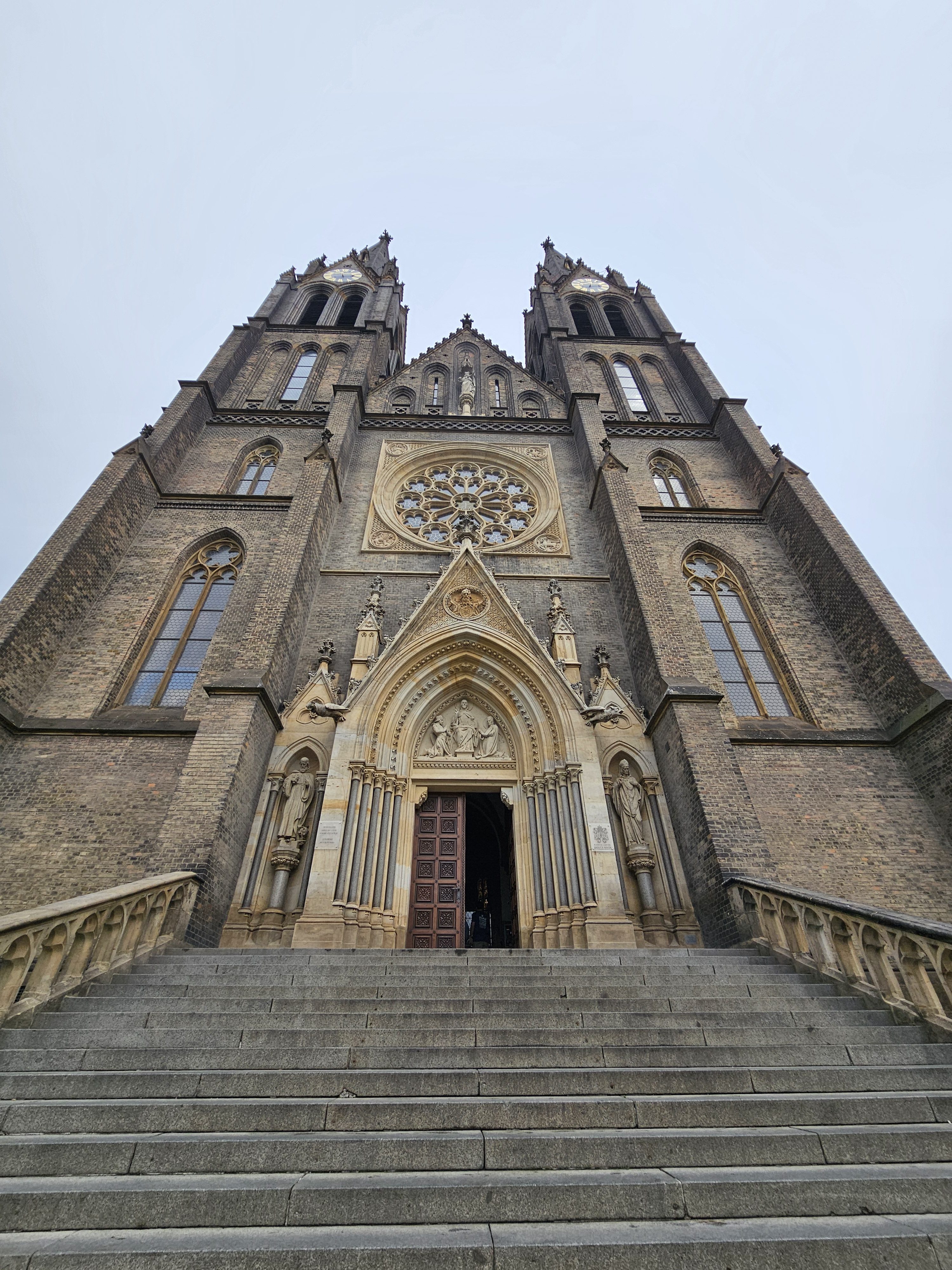 The front exterior of the Basilica of St. Ludmila (Bazilika sv. Ludmily), a large Roman Catholic church in Prague, Czech Republic. The basilica is captured from a low-angle perspective. A wide set of stone steps leads up to the main entrance, which is partially open, revealing a glimpse of the interior.