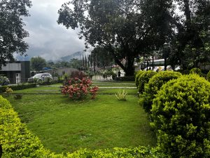 A landscaped garden with neatly trimmed bushes and flowering plants is in the foreground. In the background, there are trees and a cloudy sky, with a hilly landscape partially visible. A modern building and parked cars can also be seen.