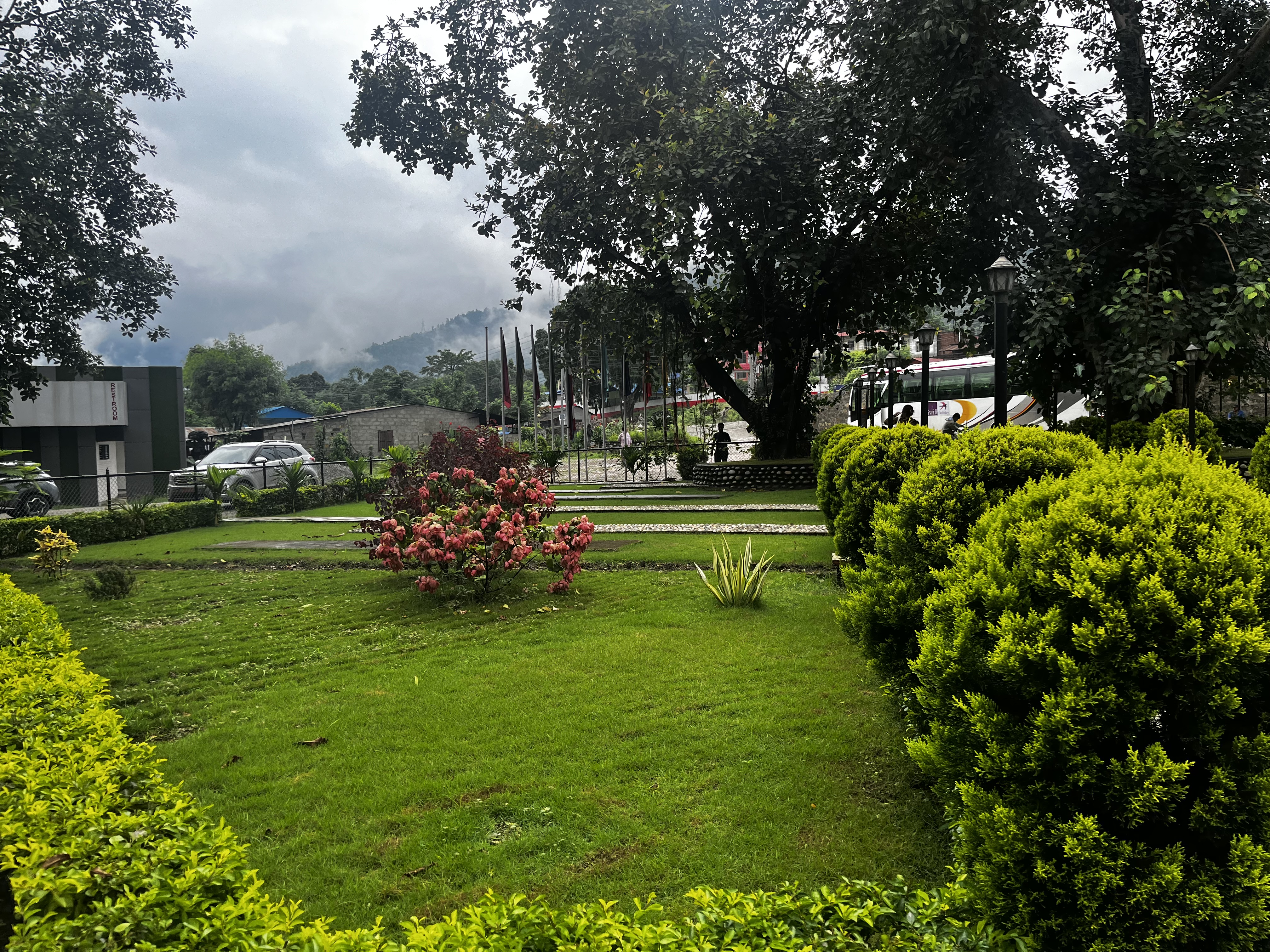 A landscaped garden with neatly trimmed bushes and flowering plants is in the foreground. In the background, there are trees and a cloudy sky, with a hilly landscape partially visible. A modern building and parked cars can also be seen.