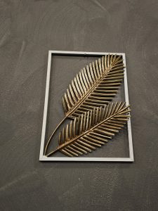 A piece of wall art featuring two golden palm leaves within a white square frame, set against a dark gray background. The leaves are metallic and textured, with detailed veins, giving them a realistic appearance. Captured from a restaurant in Turin, Italy. 
