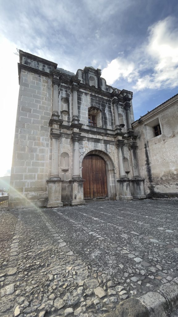 An old stone building with a wooden door and decorative columns, set against a blue sky with some clouds. The foreground features a cobblestone pathway.
