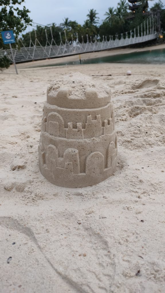 A small sandcastle with intricate details sits on a sandy beach. In the background, a rope bridge stretches across a body of water, with lush palm trees lining the shore