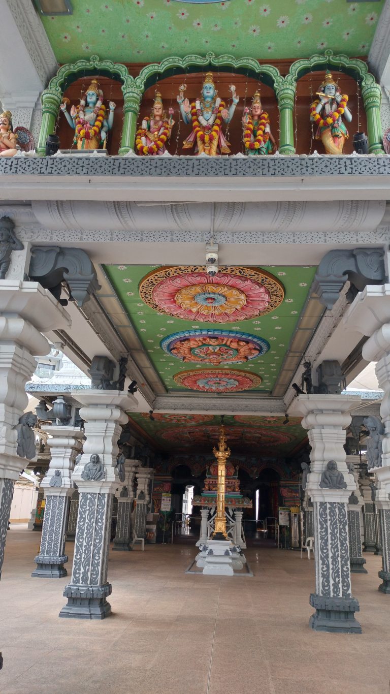 Interior of a Hindu temple featuring ornate pillars and a beautifully decorated ceiling with colorful floral patterns. Above the entrance are detailed statues of deities adorned with garlands. The pathway leads towards the inner sanctum with a golden structure at the center.