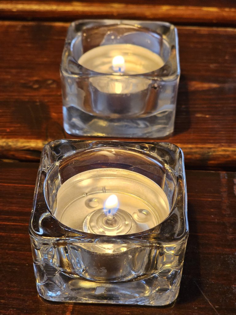 Two lit tealight candles in clear, square glass holders, set against a dark wooden surface. The candles are partially burned, with visible melted wax around the wicks. Captured from a restaurant in Prague.