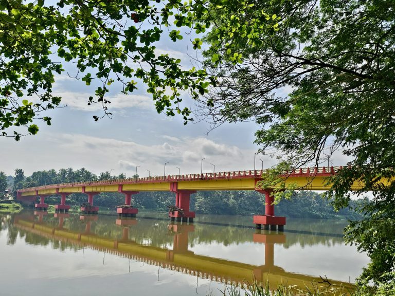 Elamaram Kadavu Bridge, a vibrant yellow and red structure, spans the Chaliyar River in Kozhikode, Kerala reflecting beautifully in the calm water below.