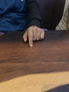 A person wearing a dark sleeve pointing at a wooden table. A partial view of another person's arm is visible on the right.