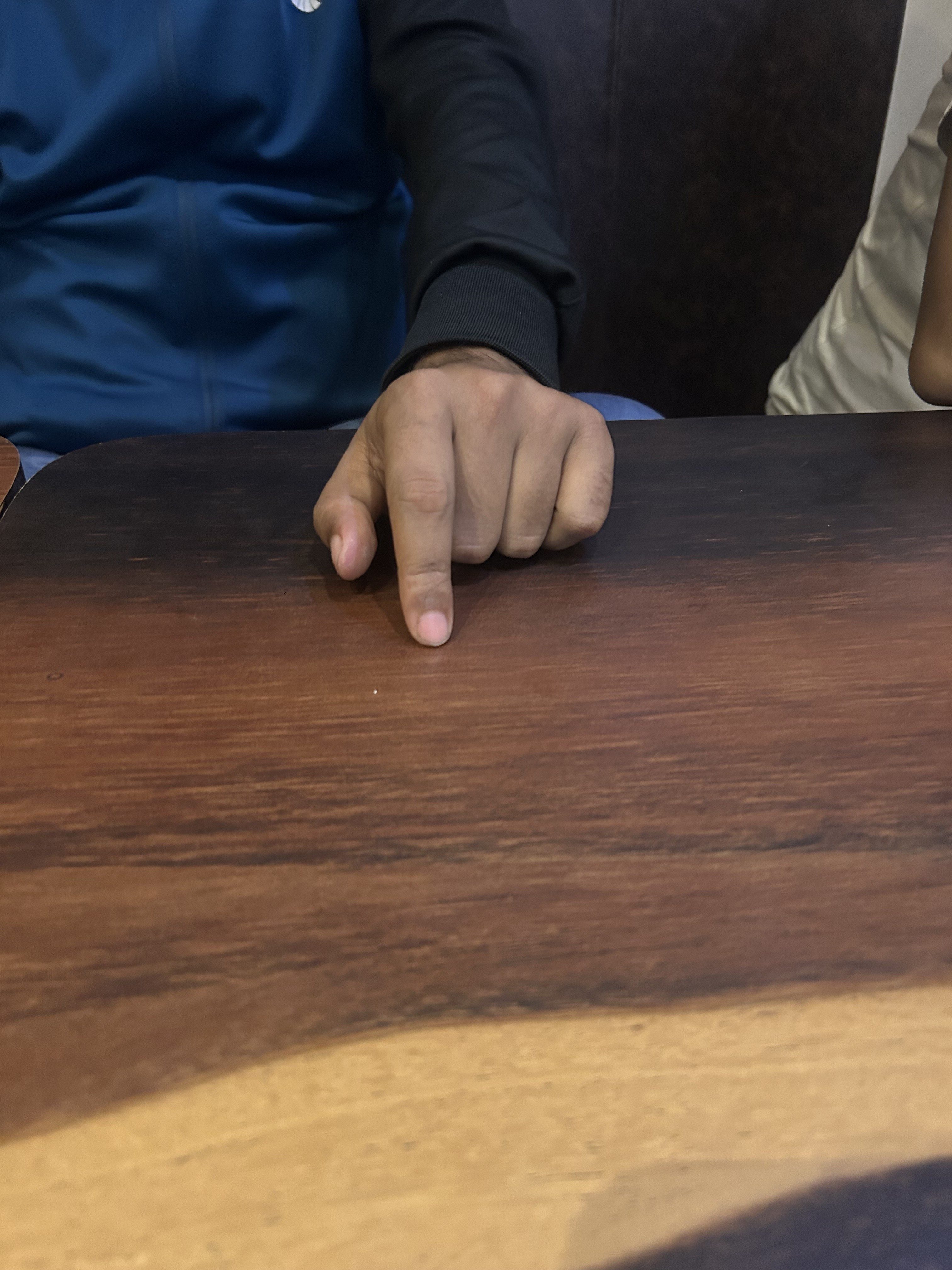 A person wearing a dark sleeve pointing at a wooden table. A partial view of another person's arm is visible on the right.