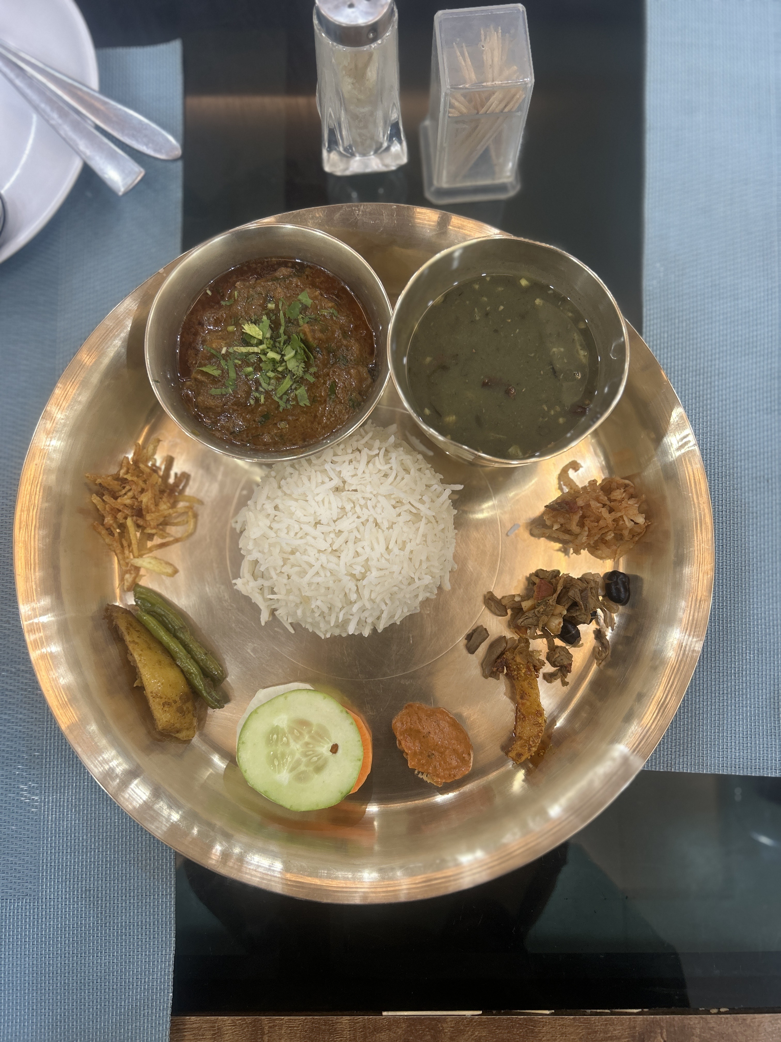 A brass plate with white rice in the center, served with two small bowls containing curry and a green vegetable dish. Accompaniments include fried strips, a green chili with a slice of fried potato, a cucumber slice with a tomato slice, a small portion of chutney, and a side of mixed vegetables.