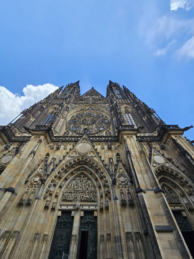 A low-angle view of the facade of St. Vitus Cathedral in Prague, with its intricate Gothic architecture reaching towards a blue sky with scattered clouds. The cathedral’s stone facade is richly decorated with carvings, arches, and sculptures, with the main entrance featuring ornate doors.