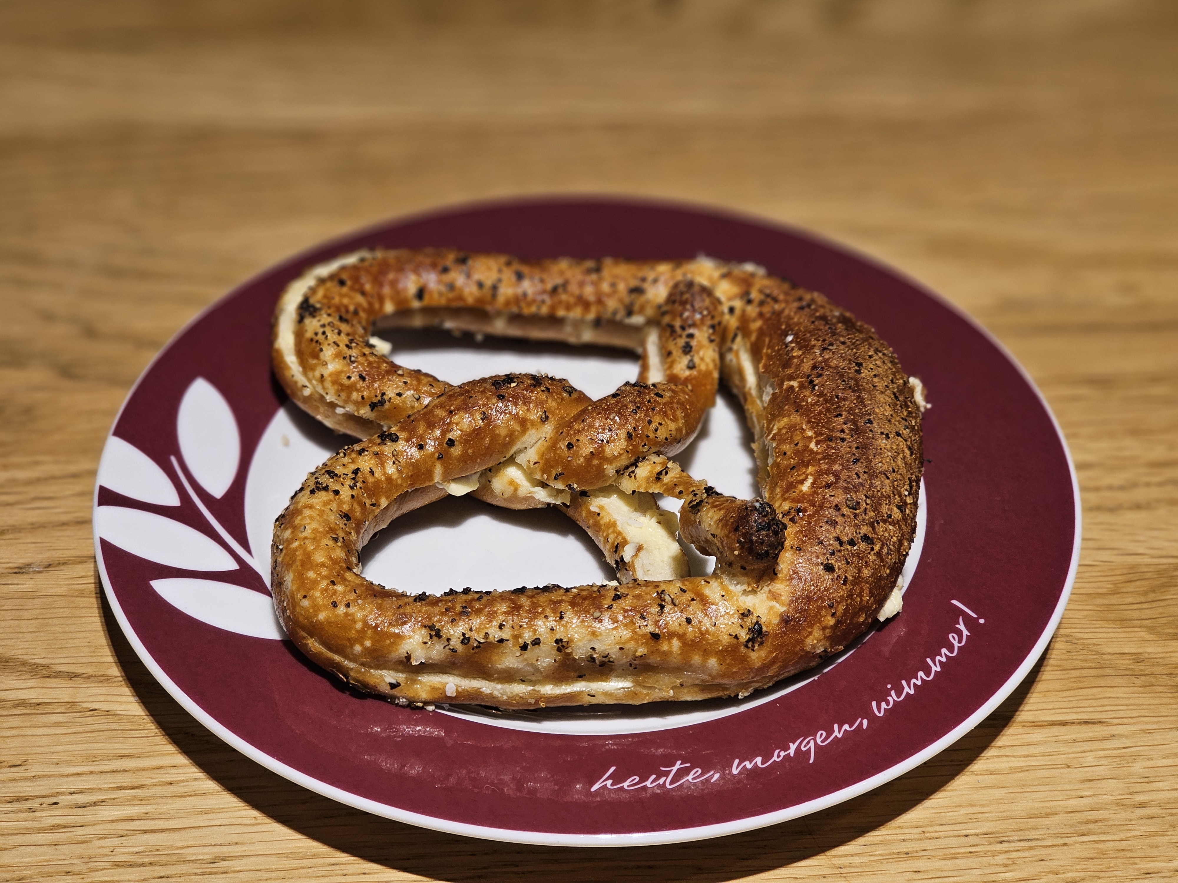 A pretzel on a plate. The pretzel is golden brown and sprinkled with black pepper, and the plate is white with a maroon rim. Captured from a restaurant in Munich, Germany.