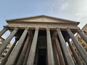 A front view of the Pantheon in Rome, Italy. The structure is an example of ancient Roman architecture, featuring a portico with large granite columns and a triangular pediment. The inscription "M AGRIPPA L F COS TERTIVM FECIT" is visible on the frieze, indicating its construction by Marcus Agrippa.