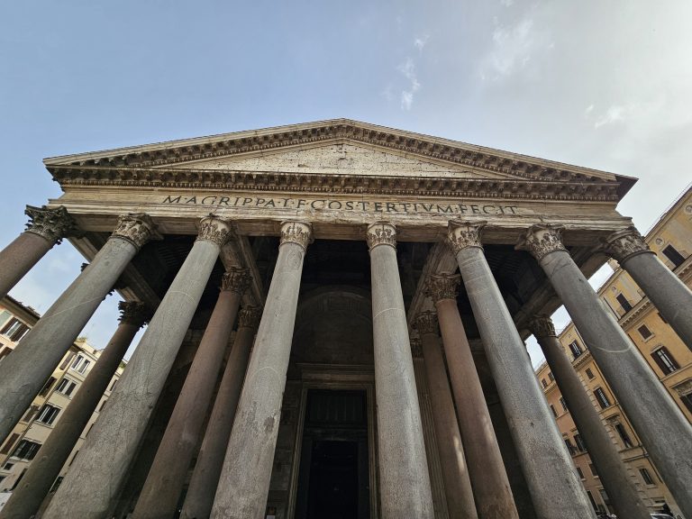 A front view of the Pantheon in Rome, Italy. The structure is an example of ancient Roman architecture, featuring a portico with large granite columns and a triangular pediment. The inscription “M AGRIPPA L F COS TERTIVM FECIT” is visible on the frieze, indicating its construction by Marcus Agrippa.