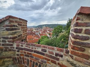 This is a cityscape of Prague, taken from Prague Castle. The view is of the city's red-tiled roofs and the green dome of St. Nicholas Church in the background. The foreground features a brick wall, which frames the view and adds depth to the composition.