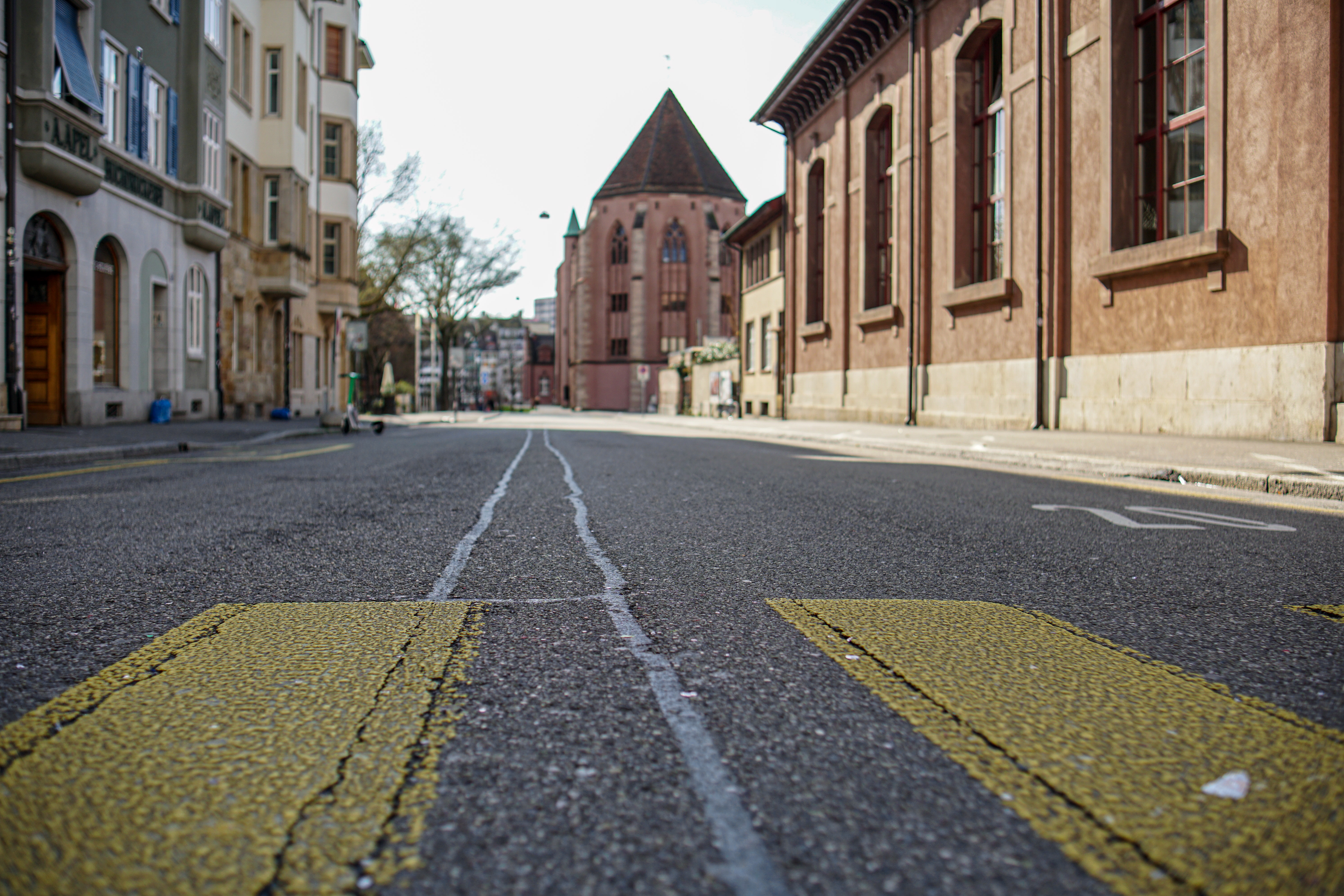 A low-angle view of an empty city street flanked by historic buildings. In the foreground, yellow road markings are visible, and a church-like structure with a pointed roof is seen in the background.