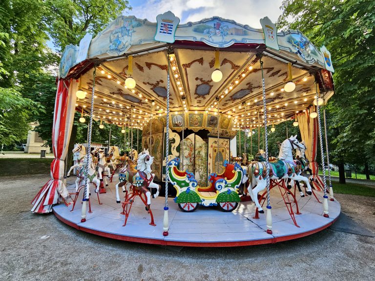 A vintage carousel in a Prague park, with classic horses, a carriage, and decorative lights. Red and white striped fabric adorns the edges, and trees in the background add to its nostalgic, old-world charm.