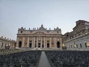 A long view of St. Peter's Basilica and St. Peter's Square in Vatican City. Rows of empty chairs are arranged in the square, suggesting preparation for an event. 