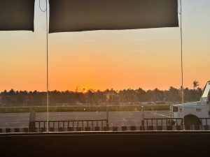 View of a sunset through a window overlooking an airport tarmac, with a silhouette of trees in the background and a portion of a ground support vehicle visible in the foreground.
