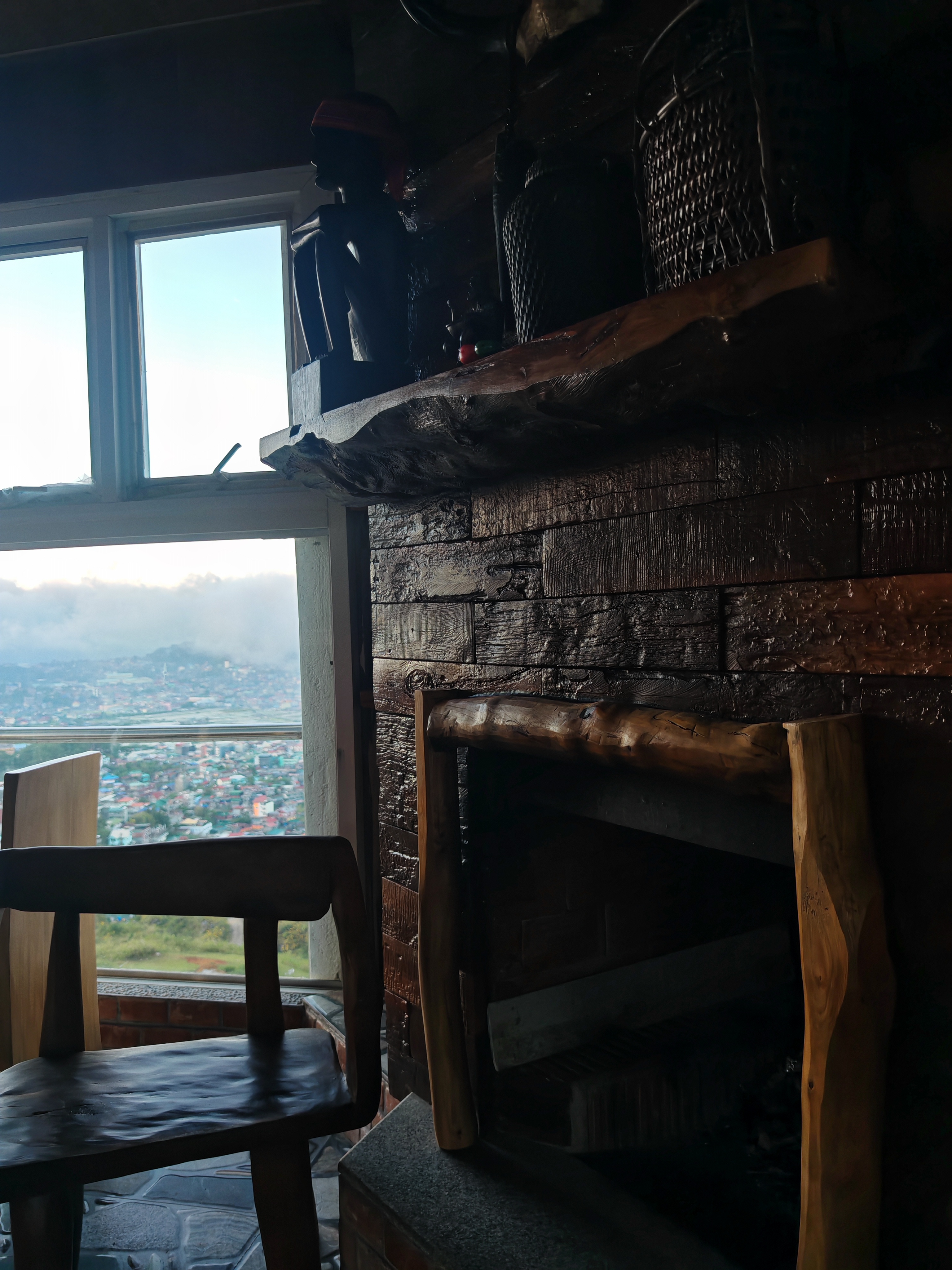 A dimly lit photo of a room with a rustic fireplace. A wooden chair sits near a window overlooking a green valley.