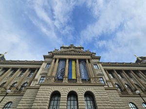 The National Museum in Prague. Banners displaying "HISTORIE HISTORY" and "PŘÍRODA NATURE," along with a blue and yellow banner in the center, hang from the columns of the building. The museum's facade is captured from a low angle, emphasizing its architectural details and the cloudy sky above.
