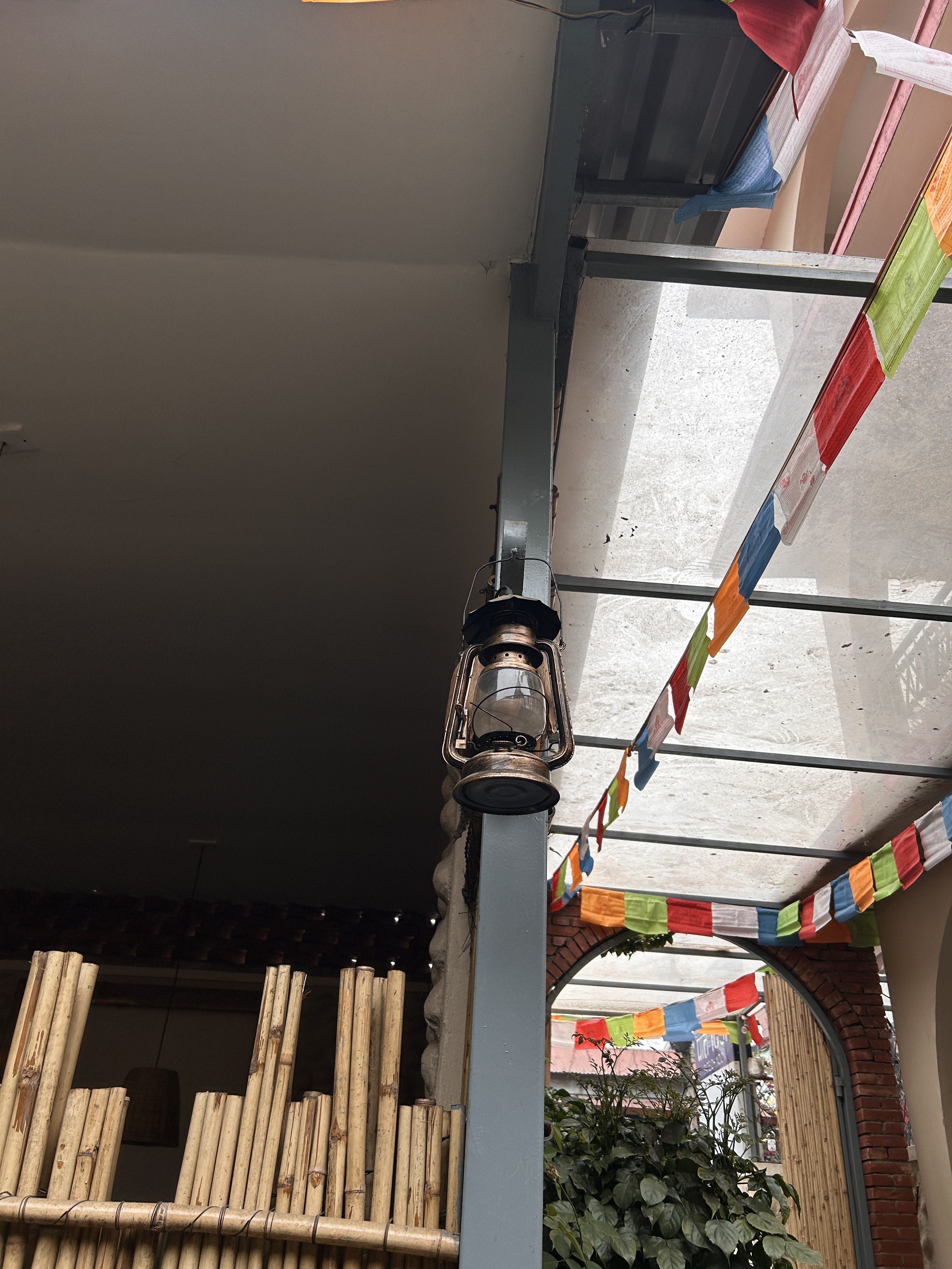 A vintage lantern is hanging from a metal beam in an outdoor area with multicolored prayer flags overhead. In the background, there is a bamboo fence and an arched entryway with greenery.