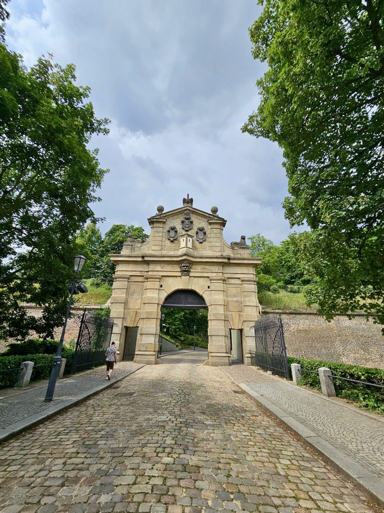 A long view of Leopold Gate (Leopoldova brána) at Vy?ehrad in Prague. The gate, constructed from light-colored stone, features a large arched opening and decorative elements at the top, including coats of arms and a sculpted face. The gate is flanked by sections of a stone wall and is accessible via a cobblestone path.