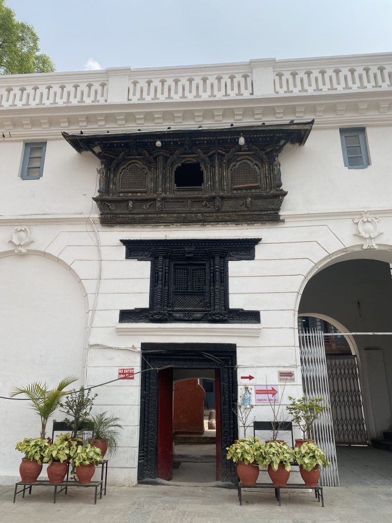 A historical building facade with intricate black woodwork above a doorway. The white wall features a central entrance flanked by potted plants on either side.