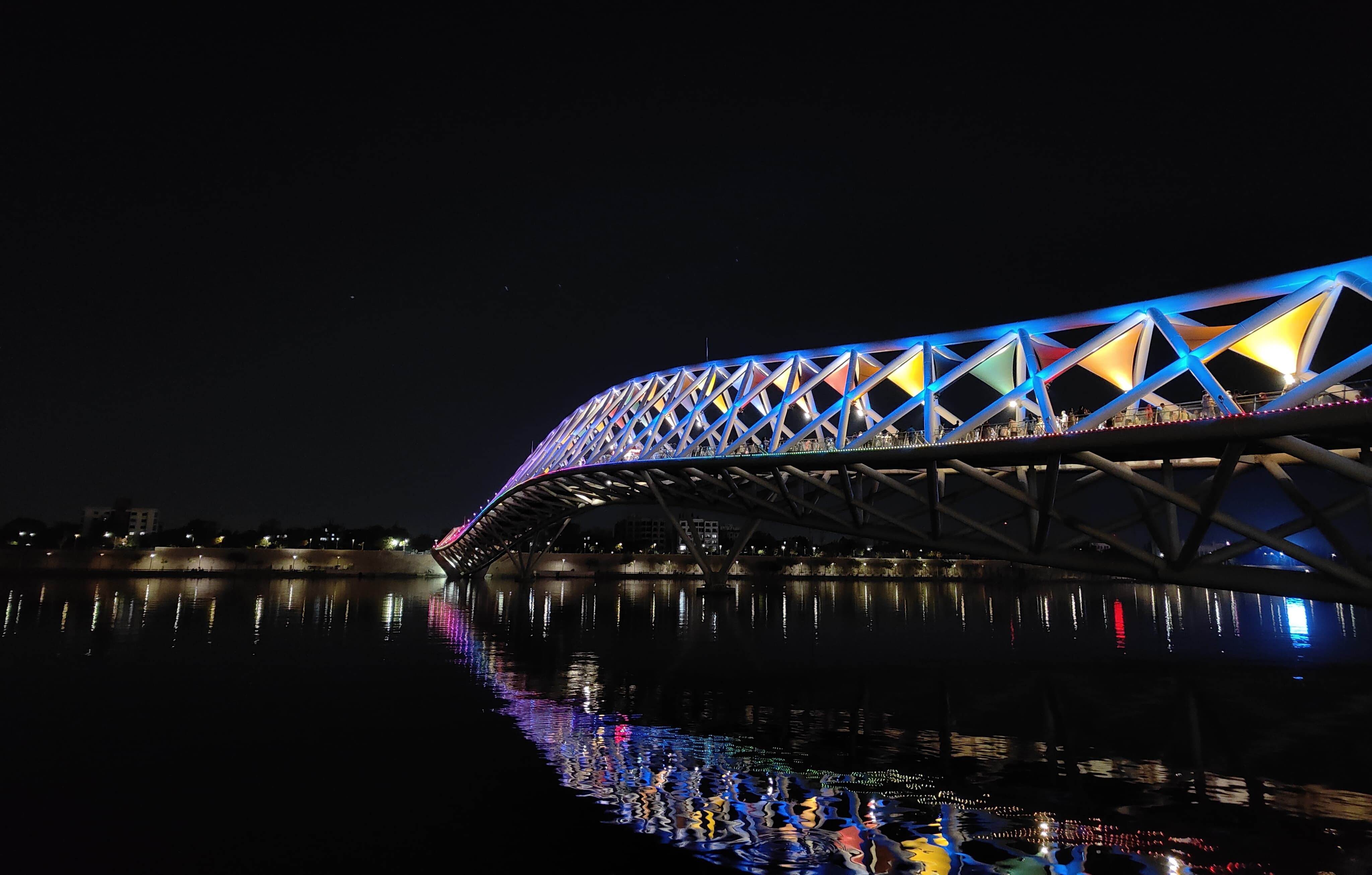 A brightly lit pedestrian bridge with a lattice structure reflects on calm water at night. Colorful lights and a dark sky enhance its striking look.