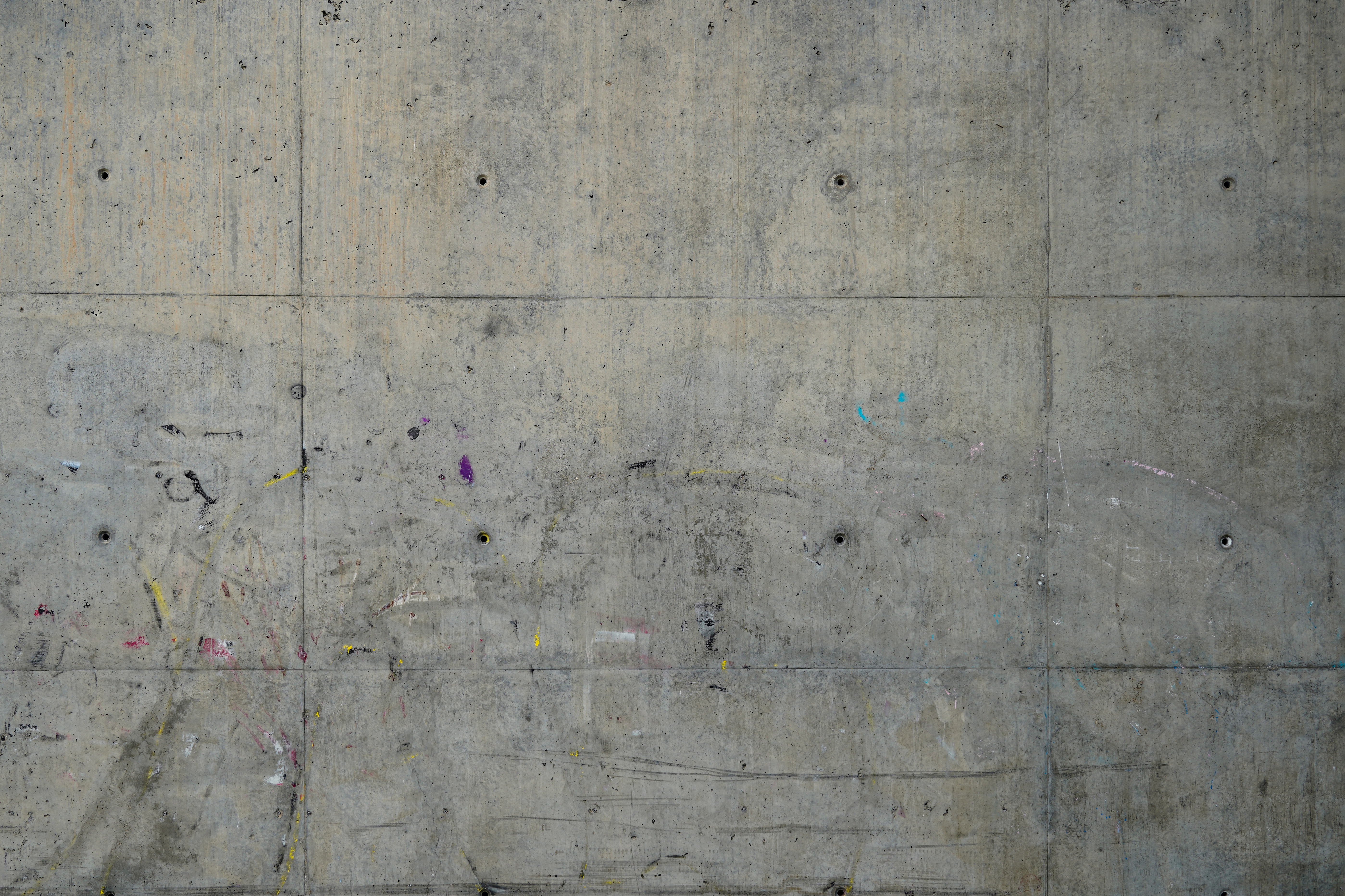 A textured concrete wall with visible seams and circular indentations, featuring faint and faded marks of colored paint smudges in various hues, such as yellow, pink, and blue.