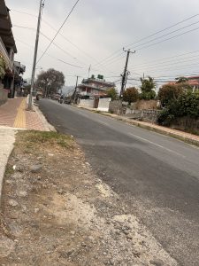 A street scene featuring a paved road lined by a sidewalk with a tactile path, utility poles with overhead wires on the right, and a few motorcycles in the distance