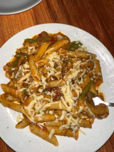 A plate of penne pasta topped with melted cheese and garnished with mixed vegetables and herbs is served on a white dish against an orange tablecloth background.
