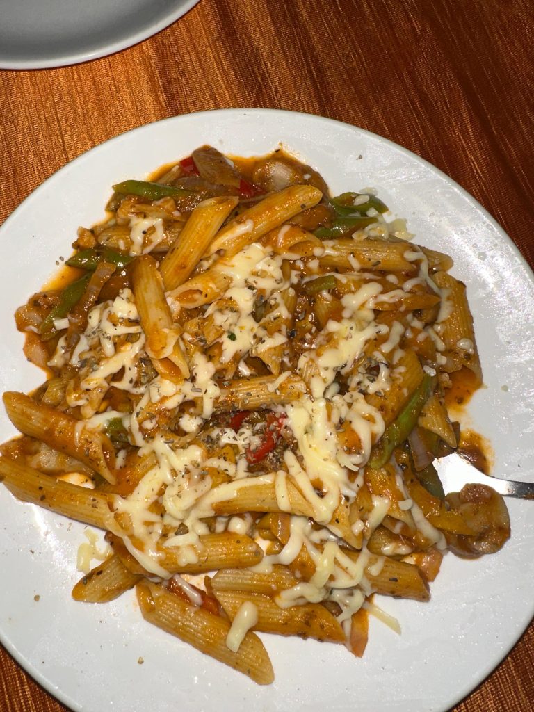 A plate of penne pasta topped with melted cheese and garnished with mixed vegetables and herbs is served on a white dish against an orange tablecloth background.