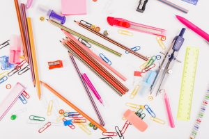 A colorful assortment of stationery items scattered on a white surface, including pencils, pens, paperclips, pushpins, erasers, rulers, a compass, a pencil sharpener, sticky notes, and a utility knife.