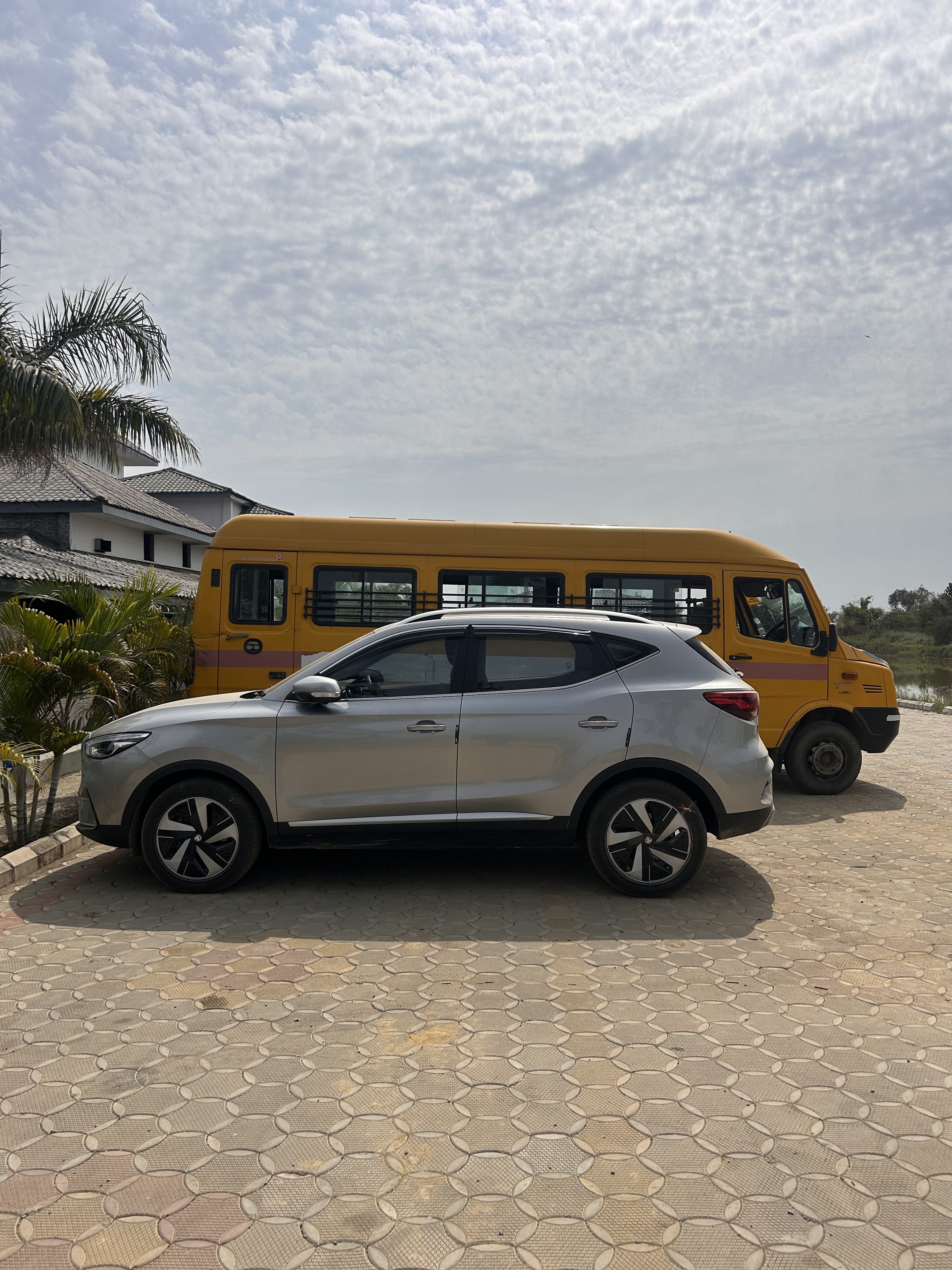 A silver SUV is parked beside a yellow school bus on a paved driveway. A palm tree and a house with a tiled roof are visible in the background under a partly cloudy sky.